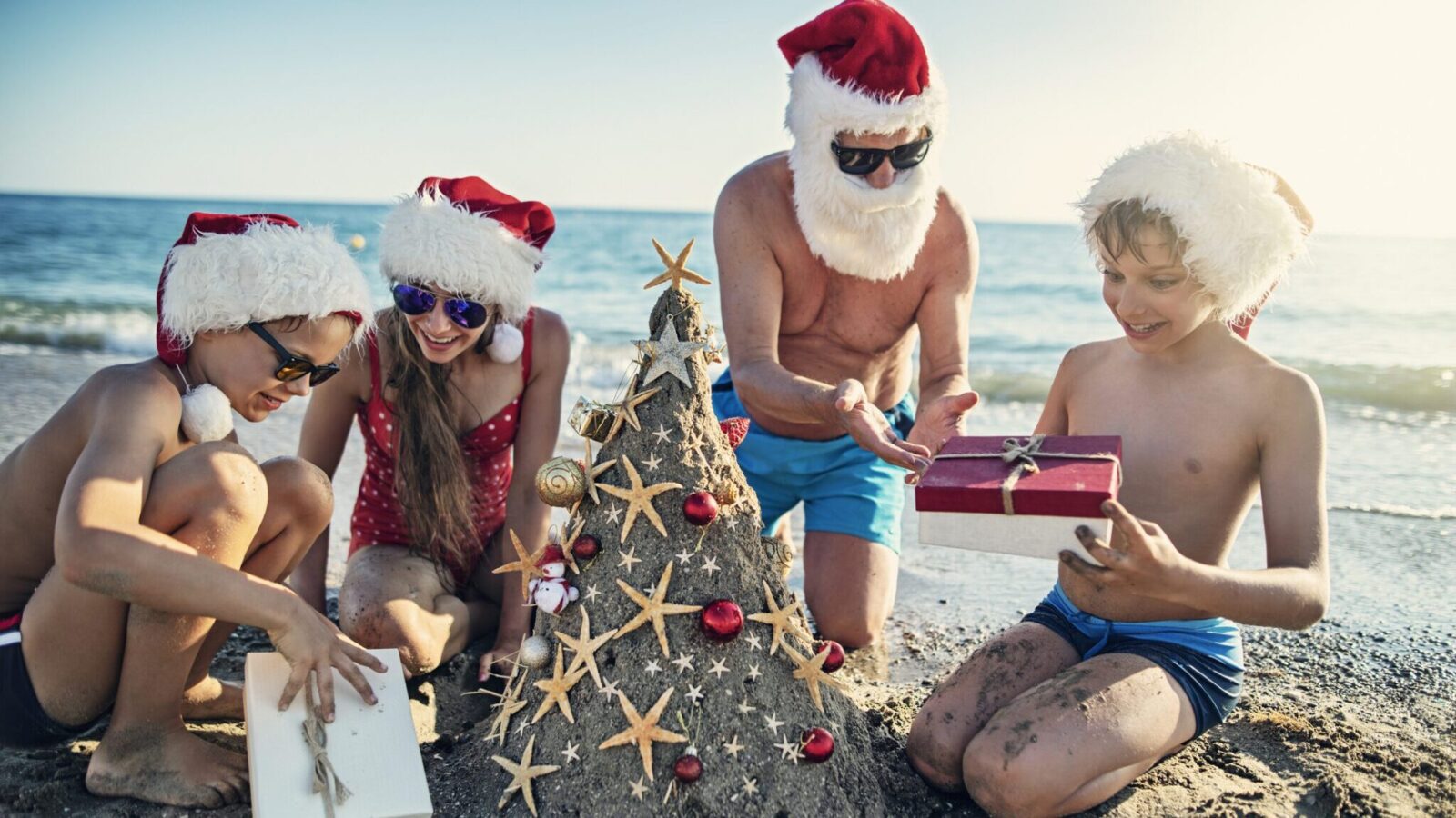 Father and three children opening gifts on the beach by a and Christmas tree.