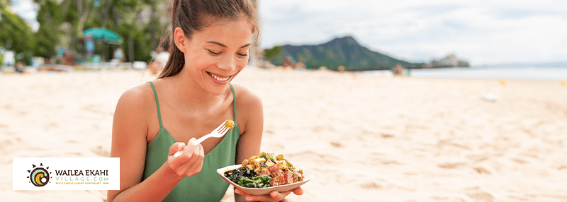 Woman on Maui beach eating a poke bowl containing raw salmon and other bright and fresh ingredients.