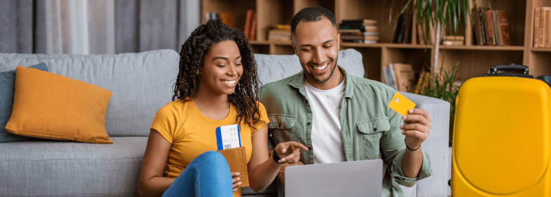Man and woman smiling at laptop and holding plane tickets and credit card with suitcase nearby